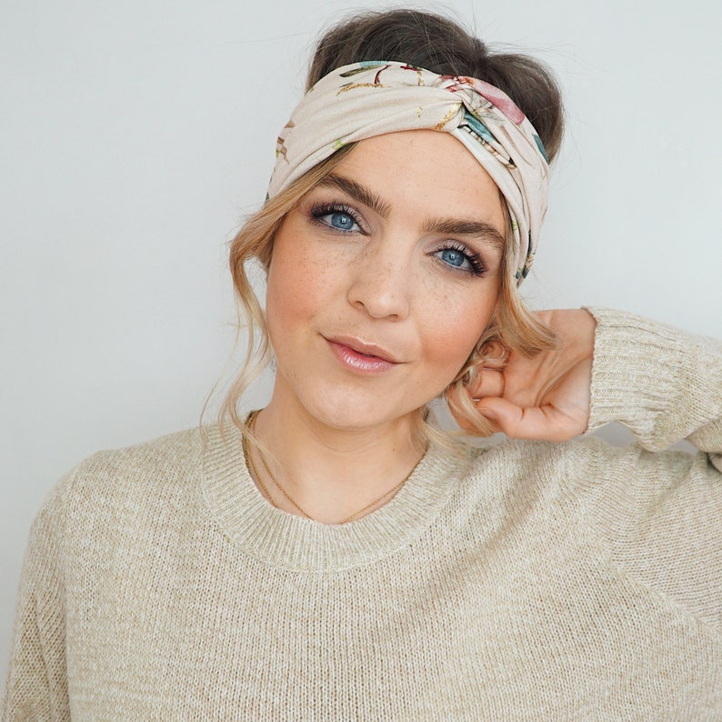K&ouml;nnte beinhalten: Ein beiges Stoffstirnband mit floralem Muster. Das Stirnband ist um den Kopf gewickelt. Die Person tr&auml;gt einen beigen Pullover. Das Haar ist zu einem Dutt frisiert.