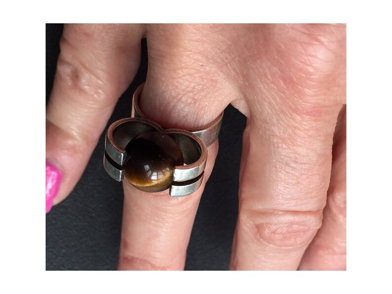 May include: A silver ring featuring a large, round, brown tiger's eye stone set in the centre. The ring has a distinctive, modern design with two bands that intersect.