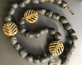 May include: A necklace featuring dark gray, rounded beads and three circular, gold-colored pendants with a leaf-like design. The necklace has a clasp and is displayed on a dark surface. The beads appear to be approximately 1 cm in diameter.