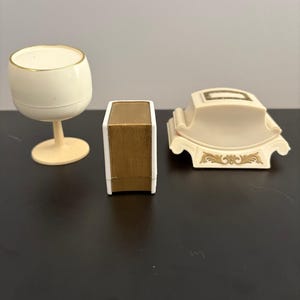 May include: Three vintage decorative items: a cream-colored wine glass with a gold rim, a small box with a gold-colored top, and a cream-colored decorative piece with gold accents. The items are arranged on a dark surface.