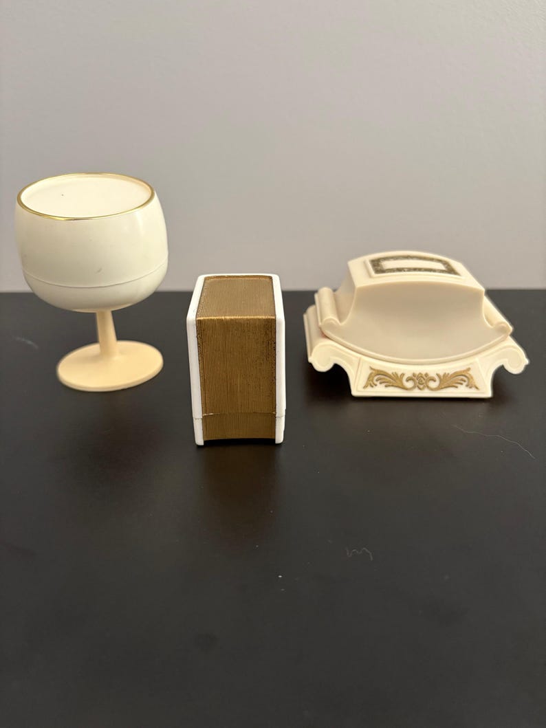 May include: Three vintage decorative items: a cream-colored wine glass with a gold rim, a small box with a gold-colored top, and a cream-colored decorative piece with gold accents. The items are arranged on a dark surface.