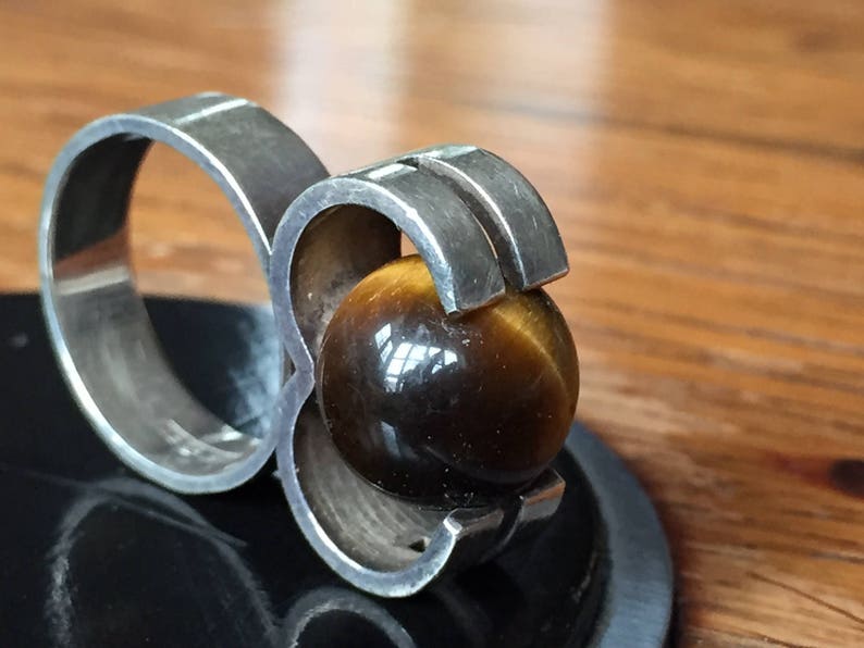 May include: A silver ring featuring a large, round, brown tiger's eye gemstone set in a geometric design.