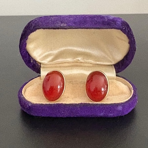 May include: A pair of oval, red gemstone earrings with gold-colored settings, displayed in an open, purple velvet jewelry box with a tan satin interior. The earrings are a classic accessory.