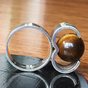 May include: A silver ring featuring a large, round tiger's eye stone set in the centre. The ring has a distinctive design with two bands that wrap around the stone.
