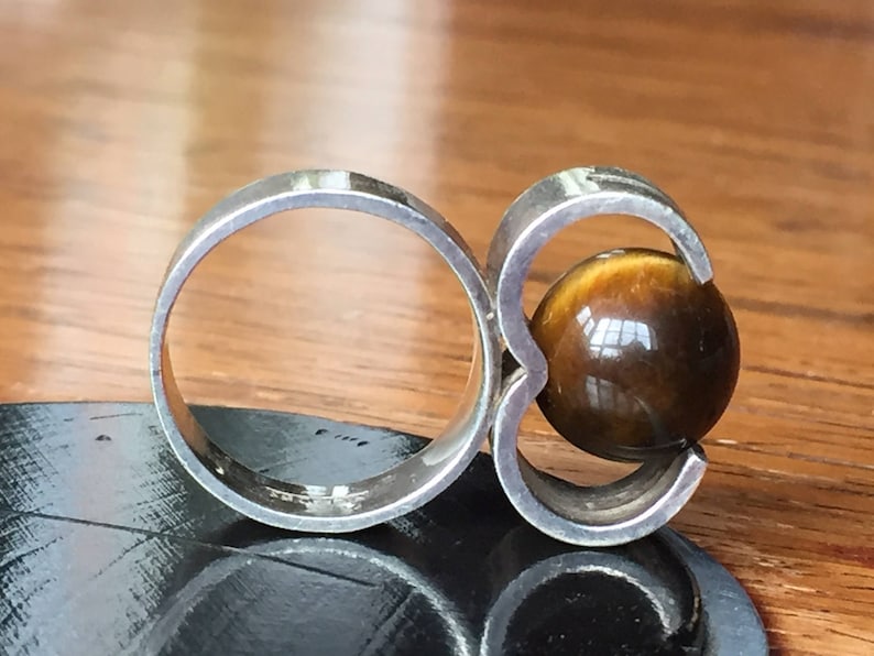 May include: A silver ring featuring a large, round tiger's eye stone set in the centre. The ring has a distinctive design with two bands that wrap around the stone.