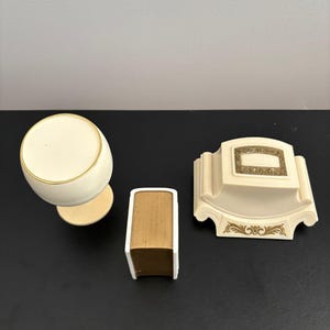 May include: Ivory-colored vanity set featuring a round-topped container, a small book-shaped item, and a decorative soap dish. The set has gold trim and accents, and is set against a dark background.