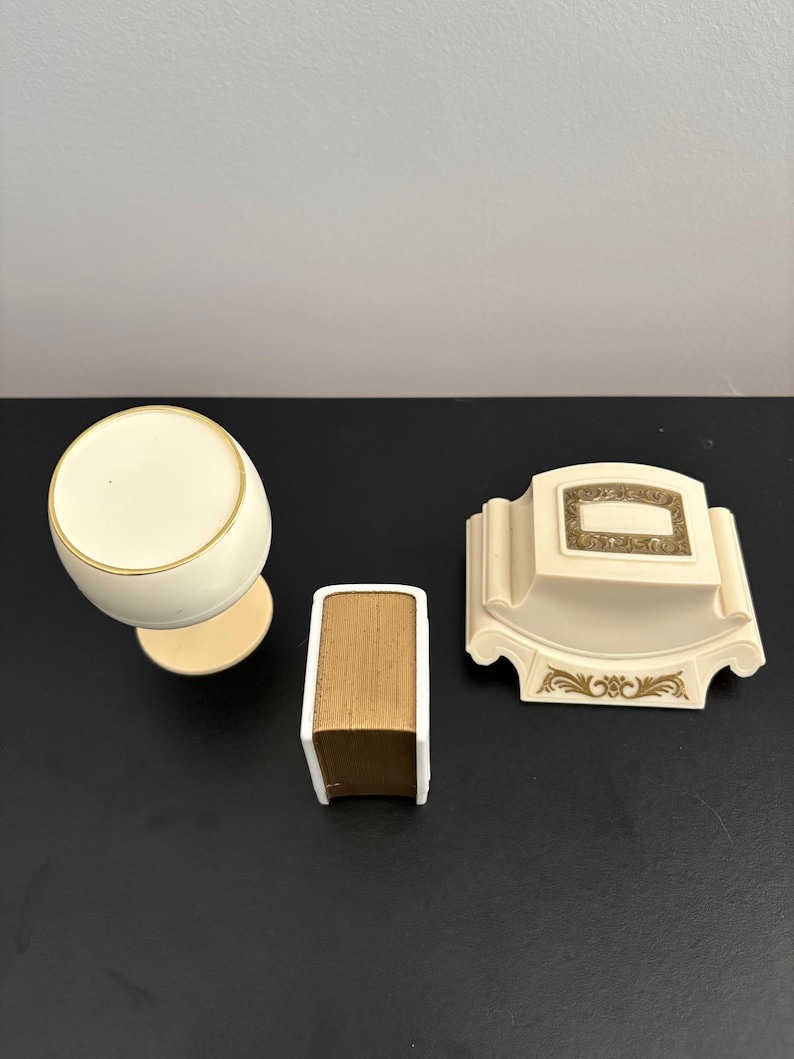 May include: Ivory-colored vanity set featuring a round-topped container, a small book-shaped item, and a decorative soap dish. The set has gold trim and accents, and is set against a dark background.