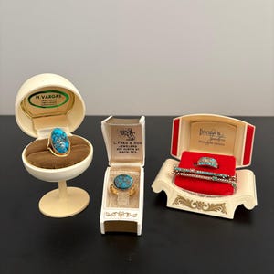 May include: An assortment of vintage jewelry pieces, including rings and a bracelet, displayed in their original boxes. The rings feature turquoise stones set in gold-toned metal. The boxes are cream and red, with gold accents and text.