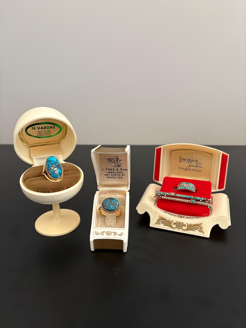 May include: An assortment of vintage jewelry pieces, including rings and a bracelet, displayed in their original boxes. The rings feature turquoise stones set in gold-toned metal. The boxes are cream and red, with gold accents and text.