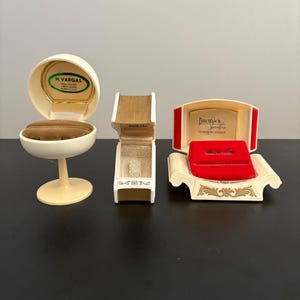 May include: Three vintage jewelry boxes in cream and gold tones. One is egg-shaped, another is rectangular, and the third is a sofa-shaped box with a red interior. The boxes have text that reads "H. Vargas", "Waco, Tex", and "Dalton's Jewelers".