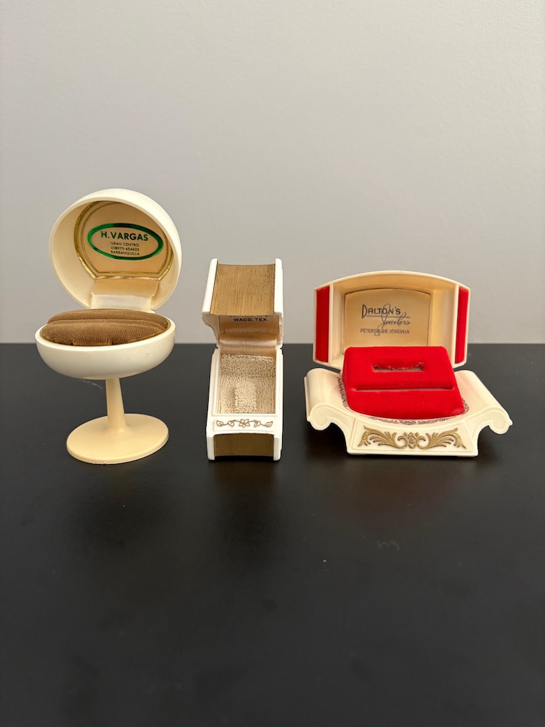 May include: Three vintage jewelry boxes in cream and gold tones. One is egg-shaped, another is rectangular, and the third is a sofa-shaped box with a red interior. The boxes have text that reads "H. Vargas", "Waco, Tex", and "Dalton's Jewelers".
