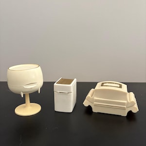 May include: Three ivory-colored decorative items: a wine glass, a small box with a gold top, and a miniature sofa. The wine glass has a rounded bowl and a pedestal base. The box is rectangular. The sofa has curved arms.