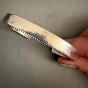 May include: A polished silver-tone cuff bracelet with a smooth, wide band and an open back design. The metal surface features a reflective finish with subtle variations in tone, showcasing a simple, unadorned, and continuous curved structure.