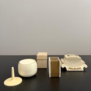 May include: A collection of vintage vanity items, including a small, off-white container with a gold rim, a small box, a rectangular box with gold accents, a decorative soap dish, and a small stand.