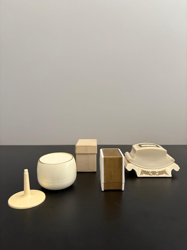 May include: A collection of vintage vanity items, including a small, off-white container with a gold rim, a small box, a rectangular box with gold accents, a decorative soap dish, and a small stand.