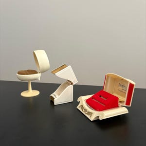 May include: Three vintage jewelry boxes in cream and gold tones. One is a small chair, another is a hinged box, and the third is an open box with a red velvet insert. The third box has the text "Dalton's" inside.