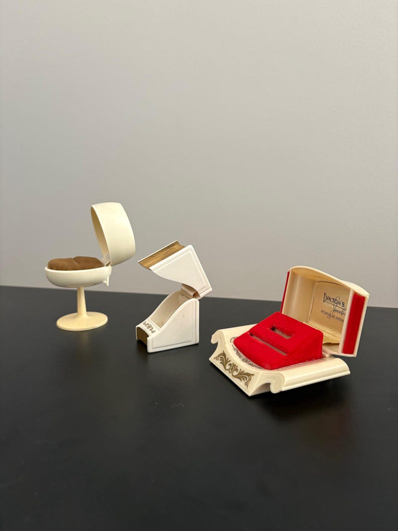 May include: Three vintage jewelry boxes in cream and gold tones. One is a small chair, another is a hinged box, and the third is an open box with a red velvet insert. The third box has the text "Dalton's" inside.
