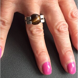 May include: A silver ring featuring a brown tiger's eye stone set in the centre. The ring has a geometric design with two silver bands on either side of the stone.
