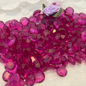 Op de afbeelding: Een grote stapel magenta gekleurde gefacetteerde glazen edelstenen. De edelstenen zijn verspreid over een wit oppervlak.