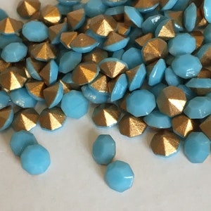 May include: A pile of turquoise blue glass rhinestones with gold accents. The rhinestones are scattered on a white surface.