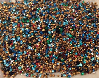 May include: A large assortment of colorful rhinestones and gold-toned studs. The rhinestones are in various shades of blue, green, red, and clear. The studs are small and have a pointed top.