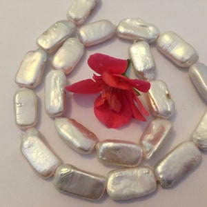 May include: A strand of white baroque freshwater pearls arranged in a spiral shape. The pearls are rectangular and have a slightly iridescent sheen. A pink flower is in the centre of the spiral.