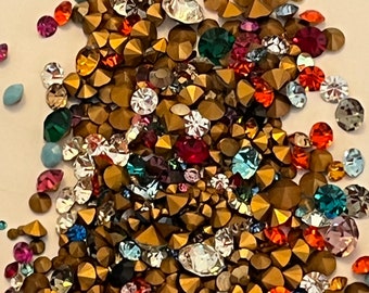 May include: A large assortment of colorful rhinestones, including gold, silver, red, blue, green, and purple. The rhinestones are scattered on a white surface.