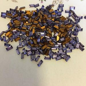 Peut inclure: Un grand tas de strass rectangulaires en verre violet et or. Les strass sont éparpillés sur une surface blanche.
