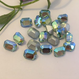 Könnte beinhalten: Eine Gruppe von regenbogenfarbenen, rechteckigen Strasssteinen in Grün und Blau. Die Strasssteine haben ein facettierendes Design und sind auf einer weißen Oberfläche verstreut.