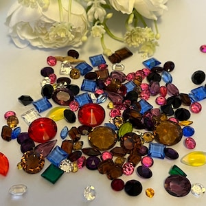 May include: A selection of multi-coloured gemstones in diverse shapes, including round, square, and teardrop. The gems are in shades of red, blue, pink, brown, and yellow. White flowers are in the background.