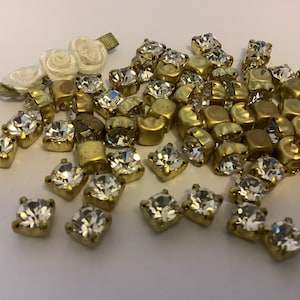 May include: A pile of clear glass rhinestones with gold settings. The rhinestones are scattered on a white surface.