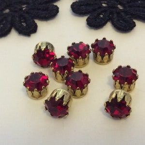 Peut inclure: Huit embellissements en strass rouge avec des montures dorées. Les strass sont ronds et ont un design facetté. Les montures sont ornées et ont une forme de couronne.