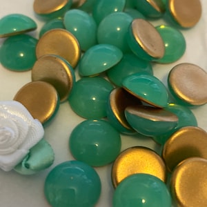 May include: A collection of round, green cabochons with gold backing. The cabochons are scattered on a white surface.