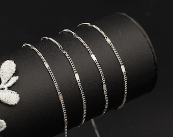 May include: Close-up of several delicate silver necklaces with a chain and rectangular bar design. The necklaces are draped over a black cylindrical display stand. The background is a solid black.