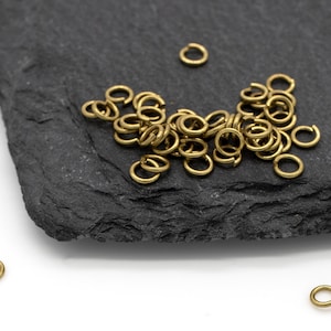 May include: A selection of small, open brass jump rings scattered on a dark grey slate surface and a white background. The jump rings are circular with a small opening, used for jewellery making and crafts.