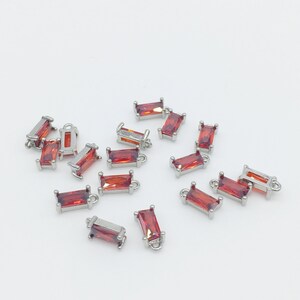 May include: A scattering of small, silver-toned charms with red, rectangular, faceted stones. The charms have a loop at the top for attaching to jewelry.
