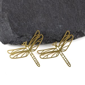 May include: Two gold-colored metal dragonfly charms with intricate cut-out details. The charms are on a black slate background.
