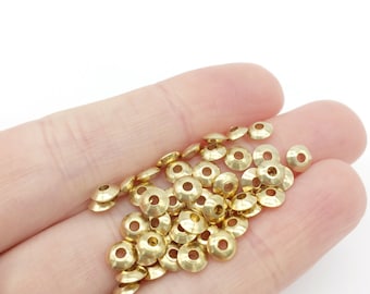 May include: A pile of gold-colored metal beads in the shape of a small, flat disc with a raised center. The beads are arranged in a loose pile in the palm of a hand.