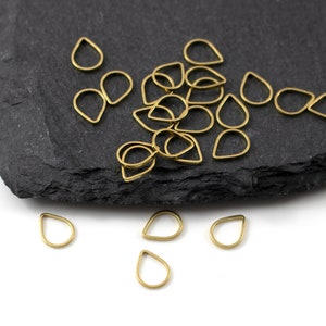 May include: A pile of gold-coloured teardrop-shaped jump rings on a black slate surface. Some of the rings are scattered on a white surface.