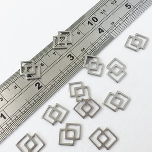 May include: A close-up of a group of silver metal double square charms. The charms are scattered on a white surface next to a ruler.