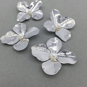 May include: Four silver flower-shaped embellishments, each with a pearl bead centre. The petals have a textured, metallic finish. The flowers are arranged on a grey surface.