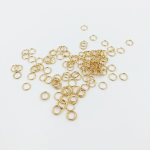May include: A scattering of small, gold-coloured jump rings on a white background. The rings are circular and appear to be metal, suitable for jewellery making or crafts. The bright lighting accentuates the metallic sheen of the rings.