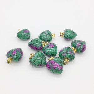 May include: A collection of heart-shaped pendants. Each pendant features a green and pink speckled stone with a gold-colored bail. The pendants are scattered on a white surface.
