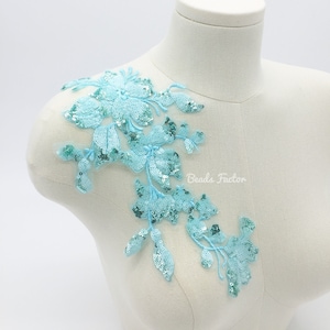 May include: A light blue floral applique with sequins, displayed on a mannequin. The applique features a delicate design of flowers and leaves, with shimmering sequins adding a touch of sparkle. The text "Beads Factor" is visible.