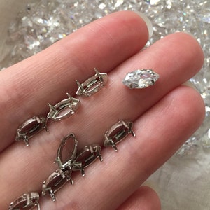 May include: A close-up of a hand holding a silver setting for a marquise-shaped gemstone. There are several other settings in the background, all of which are silver.