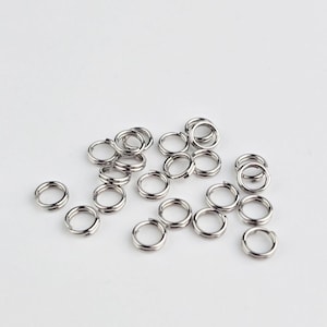May include: A scattering of small, silver split rings on a white background. These double-loop metal rings are typically used in jewellery making and crafts. The image is a close-up, showing the metallic shine of the rings. They are likely used for connecting items.