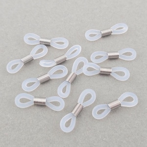 May include: A selection of clear spectacle retainers. Each retainer has a loop at each end, joined by a silver-coloured metal coil. The retainers are scattered on a light grey surface.