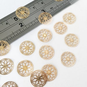 Könnte beinhalten: Eine Sammlung von 15 goldfarbenen, dekorativen Metallkreisen mit einem floralen Design. Die Kreise sind auf einer weißen Oberfläche verstreut. Ein Lineal befindet sich im Hintergrund.