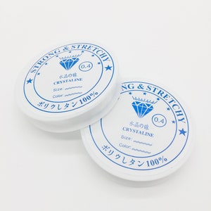 May include: Two white spools of strong and stretchy crystalline elastic cord. Each spool has a blue and white label with text including "STRONG & STRETCHY", "CRYSTALINE", and "0.4". The cord is likely used for jewellery making or crafts.