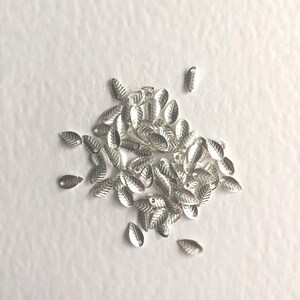以下が含まれることがあります： 上部に穴が開いた銀の葉の形をしたチャームの山。チャームは白い表面に散らばっている。